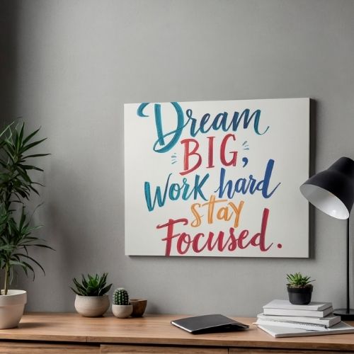 Motivational wall art with 'Dream Big, Work hard, Stay Focused' on a gray wall above a desk with plants and books.