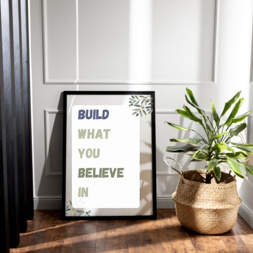 Framed motivational poster with 'Build What You Believe In' text on a wooden surface with a plant.